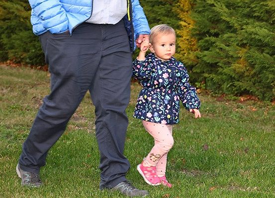 Eine Person geht mit einem kleinen Kind, das ihre Hand hält, auf dem Gras. Das Kind trägt eine Bluse mit Blumenmuster und eine pinke Hose.