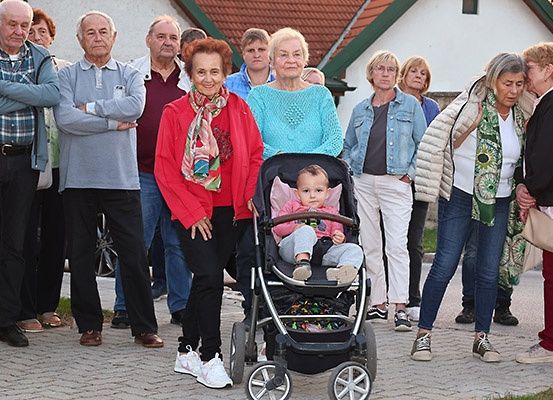 Eine Gruppe älterer Menschen steht draußen, manche mit verschränkten Armen, andere im Gespräch. Ein Baby ist in einem Kinderwagen, und zwei Frauen umarmen sich in der Nähe.