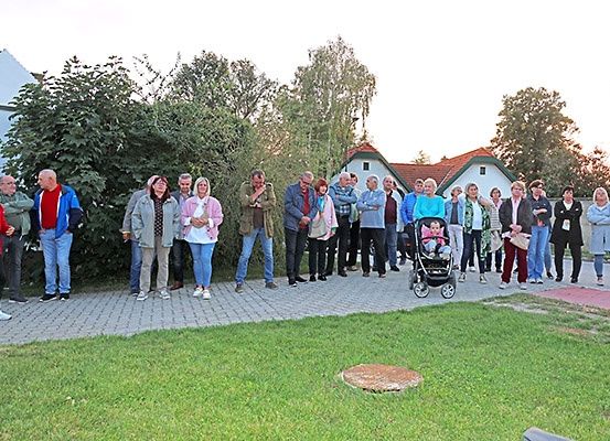 Eine Gruppe von Menschen steht in einem Außenbereich mit einem Rasen und einem gepflasterten Weg. Einige von ihnen tragen Brillen und halten einen Kinderwagen. Im Hintergrund befinden sich Häuser.