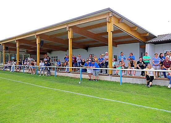 Zuschauer bei einer Sportveranstaltung unter einer überdachten Tribüne mit Holzbalken und einem Rasenfeld im Vordergrund.