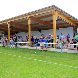 Zuschauer bei einer Sportveranstaltung unter einer überdachten Tribüne mit Holzbalken und einem Rasenfeld im Vordergrund.