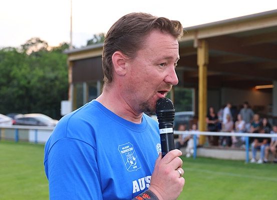Ein Mann in einem blauen T-Shirt spricht in ein Mikrofon auf einem Rasenfeld, mit einem Gebäude und Leuten im Hintergrund.