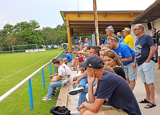 Eine Gruppe von Menschen sitzt und steht auf Tribünen bei einer Sportveranstaltung, mit unterschiedlicher Kleidung und einigen mit Brillen. Sie schauen sich das Spiel an. Die Tribünen befinden sich in der Nähe eines Feldes mit einem Tor und einem Zaun.