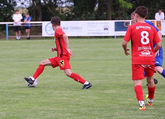 Fußballspieler in roten Trikots spielen auf einem Feld, einer kickt den Ball. Dahinter ist ein weißes Banner mit Text.
