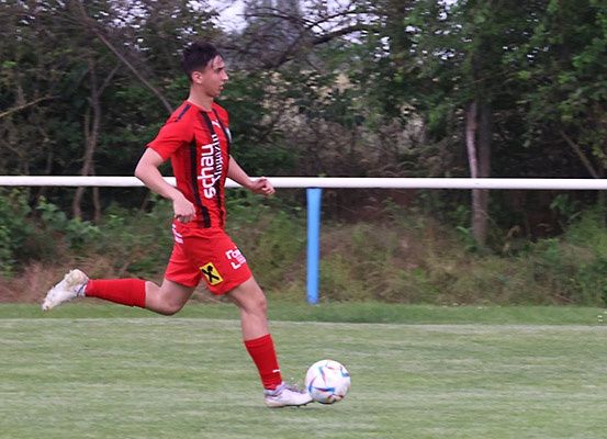 Ein Fußballspieler in einem roten Trikot läuft auf dem Feld und bereitet sich darauf vor, einen Ball zu schießen, mit einem weißen Zaun im Hintergrund.