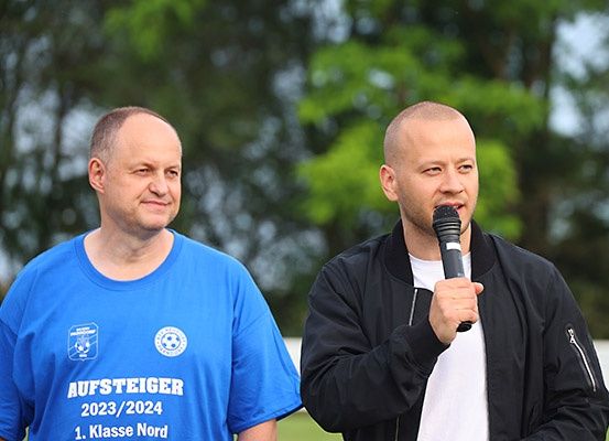 Zwei Männer stehen draußen; einer hält ein Mikrofon und der andere trägt ein blaues T-Shirt mit Text.
