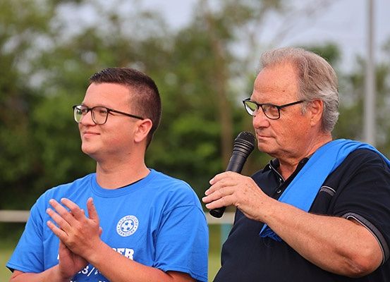 Zwei Männer stehen draußen. Einer in einem blauen Shirt klatscht, der andere in Schwarz hält ein Mikrofon. Bäume sind im Hintergrund.