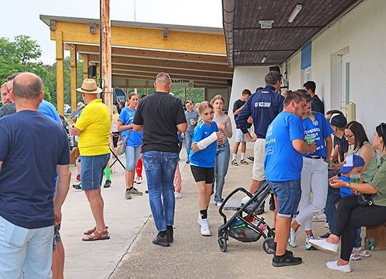 Menschen versammeln sich vor einem Gebäude, einige in blauen Shirts. Eine Frau in einem gelben Shirt steht neben einem Mann mit Hut. Ein Kinderwagen ist im Vordergrund. Bäume sind im Hintergrund sichtbar.