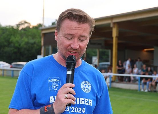 Ein Mann in einem blauen Shirt mit Logo spricht in ein Mikrofon bei einer Veranstaltung im Freien. Hinter ihm befindet sich ein Rasenbereich mit einem Metallzaun, und Personen sitzen auf einer Bank. In der Ferne steht ein Gebäude mit einem gelben Pfeiler.
