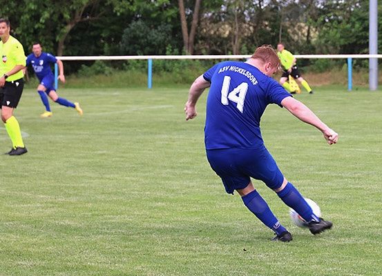 Ein Fußballspieler in Blau tritt den Ball. Zwei andere Spieler in Blau und Gelb rennen. Das Feld ist von einem weißen Zaun umgeben.