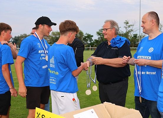 Gruppe junger Männer in blauen T-Shirts mit Medaillen, stehen auf einem Feld mit zwei Männern in Schwarz, einer mit Brille. Bäume und ein Pfahl im Hintergrund.