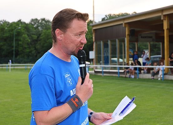 Ein Mann mit Mikrofon und Papieren spricht bei einer Outdoor-Sportveranstaltung. Im Hintergrund befinden sich ein Feld und ein Gebäude.