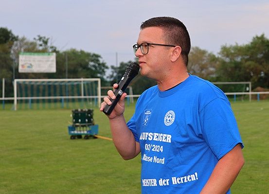 Ein Mann in einem blauen T-Shirt steht auf einem Fußballfeld, hält ein Mikrofon und spricht. Hinter ihm befindet sich ein Fußballtor und ein Rasenfeld.