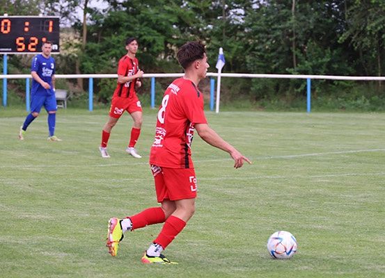 Drei Fußballspieler sind auf einem Spielfeld. Der Spieler in Rot ist dabei, den Ball zu schießen. Zwei andere Spieler rennen. Die Punktezahl ist 0:1.