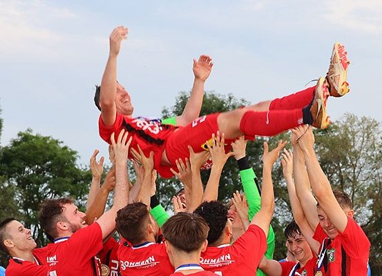 Eine Fußballmannschaft hebt einen Spieler in die Luft, um einen Sieg zu feiern. Der Spieler trägt ein rotes Trikot mit weißen Akzenten und Turnschuhen.
