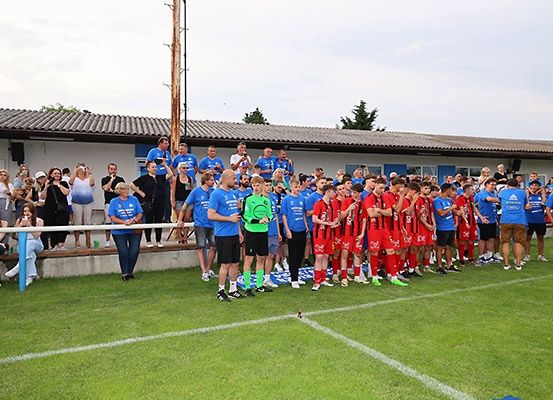 Eine Fußballmannschaft in blauen und roten Trikots steht auf dem Spielfeld, mit Zuschauern hinter einer Barriere und einem Gebäude im Hintergrund.