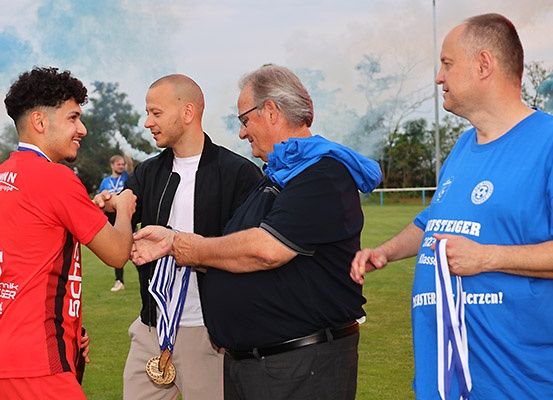 Ein Mann in einem roten Shirt schüttelt die Hand eines Mannes in einem schwarzen Jackett und mit Brille. Hinter ihnen steht ein weiterer Mann in einem blauen Shirt. Sie sind auf einem Feld mit einem unscharfen Hintergrund.