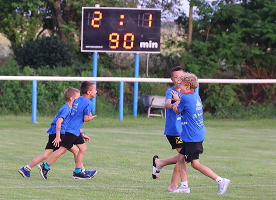 Vier Jungen in blauen Hemden und Shorts laufen auf einem Rasenfeld. Eine Anzeigetafel zeigt Punkte und Zeit. Bäume und ein Zaun sind im Hintergrund.