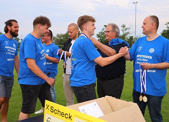 Eine Gruppe von Männern in blauen T-Shirts steht auf einem Rasenfeld, wobei einer einen Orden hält und mit einem anderen Mann die Hände schüttelt. Sie stehen vor einer Kiste.