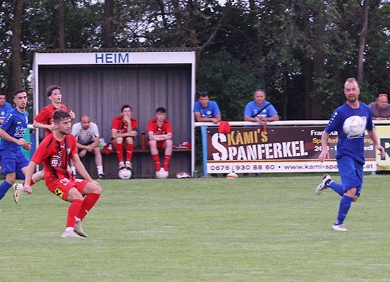 Fußballspieler auf einem Feld, einer tritt den Ball, andere sitzen auf einer Bank. Ein Banner heißt 'KAMI'S SPANFERKEL'.