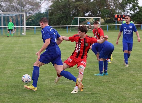 Fußballspieler in blauen und roten Uniformen spielen auf einem Rasenfeld. Der Spieler in Blau tritt den Ball, während der Spieler in Rot versucht, ihn zu blockieren. Im Hintergrund befinden sich weitere Spieler.