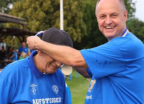 Zwei Männer in blauen T-Shirts lächeln und lachen. Einer hält ein weißes Objekt, möglicherweise ein Frisbee, in der Nähe des Kopfes des anderen Mannes. Hinter ihnen sitzen Leute an einem Tisch, und ein Gebäude mit einem Dach ist sichtbar. Bäume befinden sich im Hintergrund.