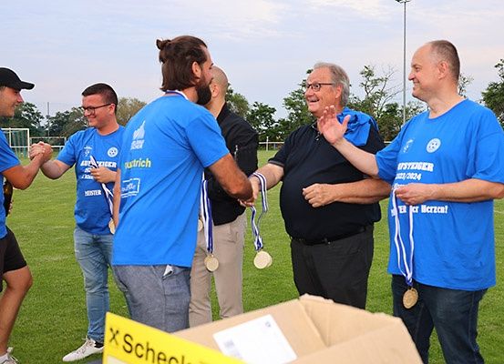 Fünf Männer in blauen T-Shirts und Medaillen stehen auf einem Feld. Ein Mann schüttelt die Hände von zwei anderen, und zwei Männer lächeln. Eine gelbe Box ist in der Nähe.