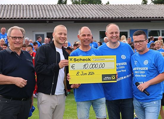 Fünf Männer in blauen T-Shirts halten einen gelben Scheck mit den Worten 'Scheck' und dem Betrag '10.000,00'. Sie stehen draußen vor einer Menschenmenge.
