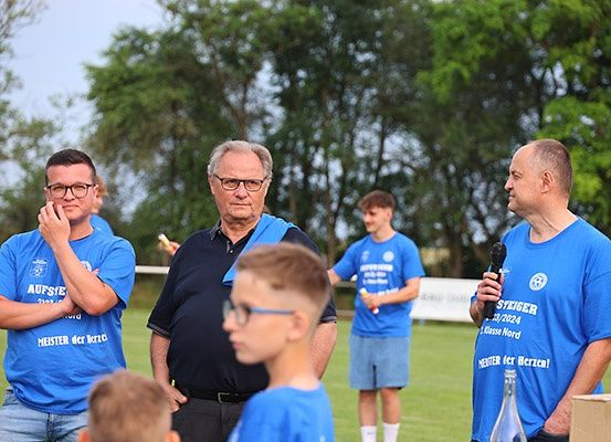 Eine Gruppe von Menschen steht auf einem Rasenfeld, einige von ihnen tragen blaue T-Shirts. Ein Mann hält ein Mikrofon, und andere schauen in verschiedene Richtungen. Hinter ihnen ist ein Banner teilweise sichtbar.