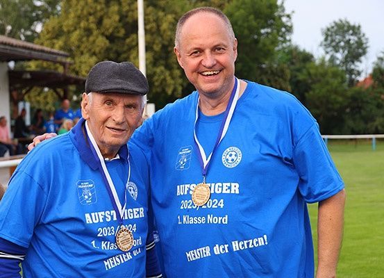 Zwei Männer stehen nah beieinander, lächeln, tragen blaue T-Shirts mit Text, halten Medaillen, einer in einem Hut, im Freien.