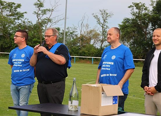 Vier Männer stehen auf einem Rasenfeld, einer hält ein Mikrofon. Sie tragen blaue T-Shirts mit weißen Logos. Ein Tisch vor ihnen hat eine Flasche und eine Pappschachtel. Der Hintergrund ist unscharf mit Bäumen.