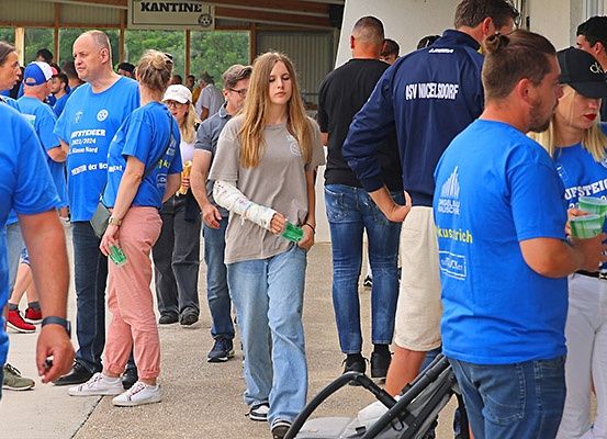 Eine Gruppe von Menschen in blauen T-Shirts steht in einer Schlange. Ein Mädchen mit einem Gips auf dem Arm schaut nach unten. Ein Mann und eine Frau schauen sich an.