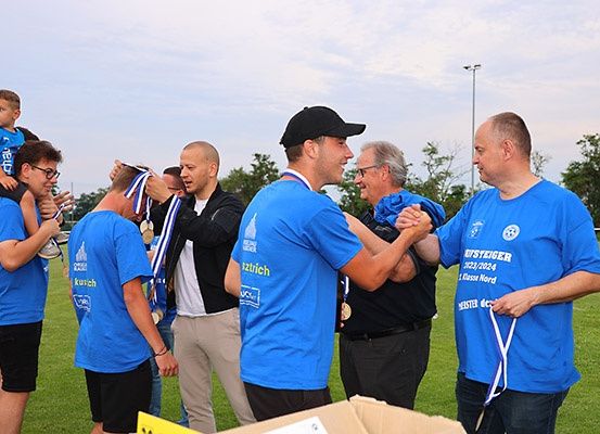 Eine Gruppe von Menschen in blauen T-Shirts feiert auf einem Rasenfeld. Sie halten Medaillen und scheinen für ein Foto zu posieren. Hinter ihnen steht ein Junge auf der Schulter eines Mannes.