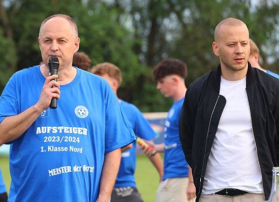 Ein Mann mit einem Mikrofon spricht zu einem anderen Mann, beide tragen blaue T-Shirts. Dahinter stehen weitere Männer in blauen T-Shirts. Im Hintergrund befinden sich Bäume und ein grasbewachsenes Feld.