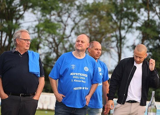 Vier Männer stehen im Freien, zwei von ihnen tragen blaue T-Shirts mit der Aufschrift 'Aufsteiger 2023/2024 1. Klasse Nord'. Der Mann auf der rechten Seite hält eine Flasche, während die anderen in ein Gespräch vertieft zu sein scheinen. Bäume und ein Zaun sind im Hintergrund zu sehen.