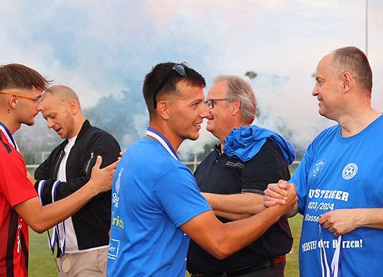 Fünf Männer in blauen und roten Trikots auf einem Feld geben sich lächelnd die Hand; zwei haben Medaillen.