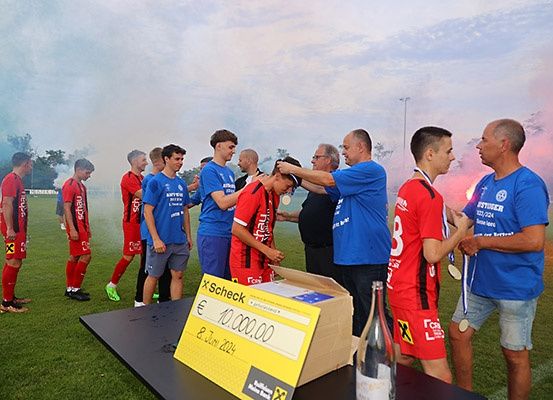 Eine Fußballmannschaft feiert auf einem Feld mit blauem Rauch und einem großen Scheck im Vordergrund. Spieler in roten und blauen Trikots stehen in einem Kreis um einen Mann in einem blauen Hemd.