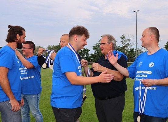 Eine Gruppe von Männern in blauen T-Shirts, einige mit Medaillen, stehen auf einem Rasenfeld. Zwei Männer in der Mitte erhalten Medaillen von den Männern an ihren Seiten. Sie scheinen einen Sieg zu feiern.