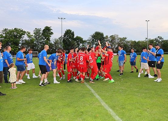 Eine Fußballmannschaft in roten Trikots feiert auf einem Feld, während Blauhemden jubeln, unter einem teilweise bewölkten Himmel. Bäume und Straßenlaternen umgeben das Gebiet.