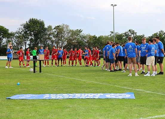 Eine Gruppe junger Fußballspieler in blauen Trikots versammelt sich auf einem Feld, mit einem Fußball auf dem Boden. Das Feld ist mit weißen Linien markiert.
