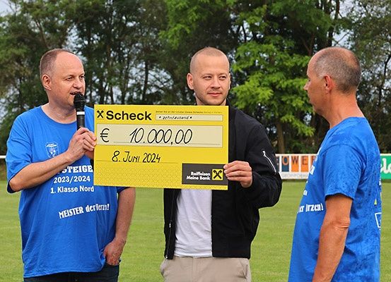 Drei Männer stehen auf einem Feld, einer hält ein Mikrofon und ein anderer einen gelben Scheck über 10.000 Euro. Der Mann mit dem Mikrofon spricht.