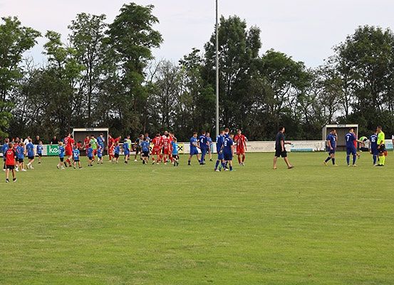 Ein Fußballspiel findet auf einem Feld statt, auf dem Spieler in blauen und roten Uniformen stehen. Einige Spieler gehen, während andere stehen. Im Hintergrund befinden sich Bäume und eine weiße Struktur.