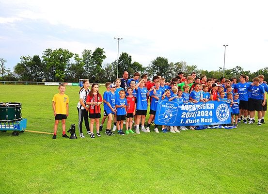 Eine Gruppe von Kindern und Erwachsenen versammelt sich auf einem Rasenfeld für eine Sportveranstaltung und hält ein blaues Banner mit Text und Zahlen.