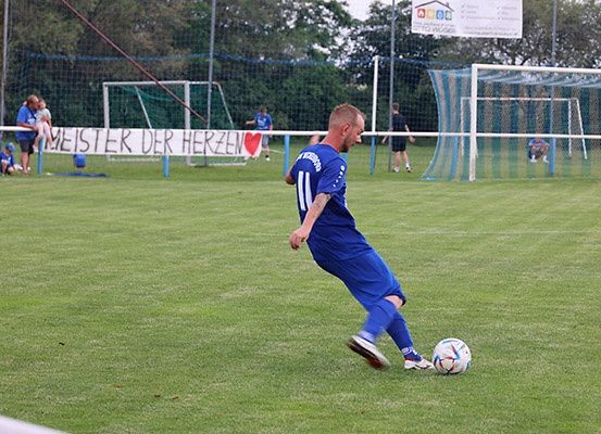 Ein Fußballspieler in einem blauen Trikot tritt einen Fußball auf einem Feld mit einem Banner, auf dem 'Meister der Herzen' steht. Im Hintergrund stehen einige Leute in der Nähe des Tores und andere Spieler sind verstreut.