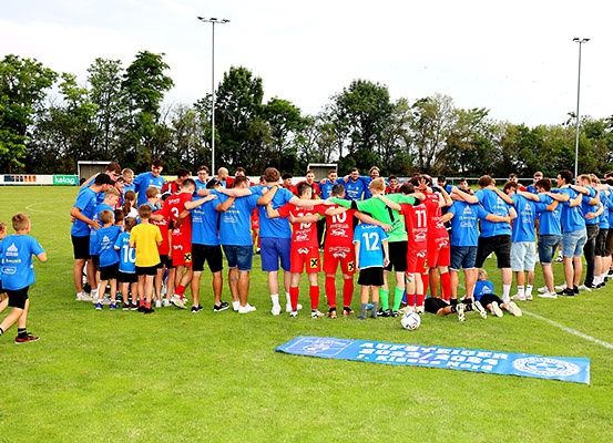 Eine Fußballmannschaft in blauen Uniformen steht im Kreis mit einem laufenden Kind. Ein Fußball liegt auf dem Boden.
