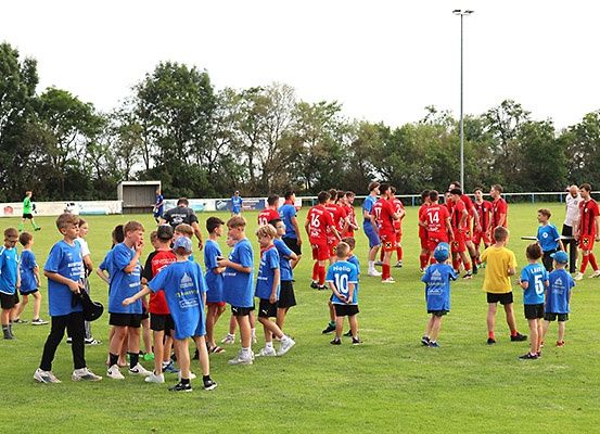 Eine Gruppe von Kindern in blauen Trikots steht auf einem Rasenfeld, wahrscheinlich einem Fußballfeld. Sie schauen eine Gruppe von Menschen in roten Trikots an. Das Feld ist von Bäumen umgeben.
