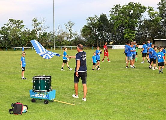 Eine Gruppe junger Fußballspieler in blauen Uniformen versammelt sich auf einem Feld. Ein Mann steht vor ihnen und hält eine Flagge. Ein Schlagzeug steht auf dem Rasen. Bäume säumen den Rand des Feldes.