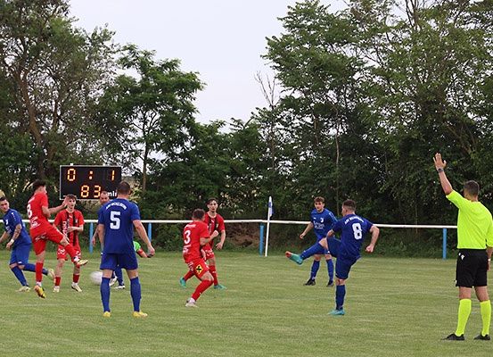 Ein Fußballspiel mit Spielern in roten und blauen Uniformen, mit einem Anzeigetafelstand von 0:1, findet auf einem grünen Spielfeld mit Bäumen im Hintergrund statt.