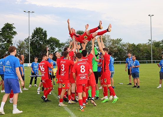 Ein Fußballteam feiert einen Sieg auf einem Feld mit Bäumen und einem bewölkten Himmel. Die Spieler tragen rote Trikots, und ein Spieler wird von anderen hochgehoben.