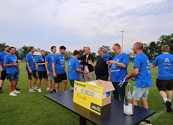 Eine Gruppe von Männern in blauen T-Shirts versammelt sich um einen Tisch auf einem Fußballfeld. Ein gelber Kasten mit der Aufschrift 'X-Scheck' und einem Wert von 10.000 Euro steht auf dem Tisch.